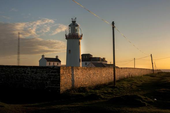 Un long mur qui délimite l’espace dans lequel se trouve un phare reçoit la lumière orangée du soleil couchant. Des poteaux télégraphiques se détachent dans un ciel presque sans nuages. 