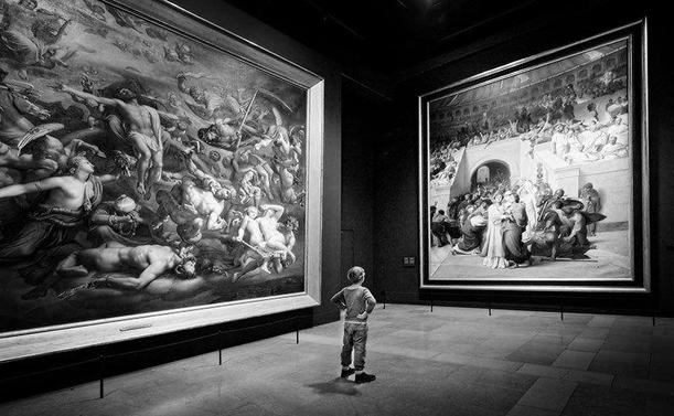 Black and white image of a small boy standing in front of a very large painting in a museum, studying it with his hands to his sides. On his left is another gigantic image.