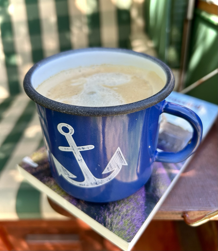 Das Bild zeigt eine blau emaillierte Tasse mit Ankermotiv, die mit einem Kaffee gefüllt ist. Die Tasse steht auf einem Buch mit einem Lavendelbild auf dem Umschlag. Im Hintergrund ist eine grün-beige gestreifte Gartenliege unscharf zu sehen. Das Bild vermittelt ein Gefühl von Ruhe und Entspannung, im Freien an einem sonnigen Tag.