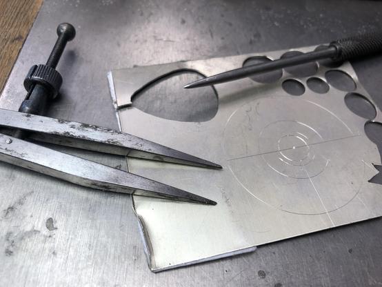 A close-up of a metalworking setup featuring a scribing compass, a pointy scriber and a marked  circular lines on a silver sheet with different cutouts, all resting on a metallic surface.