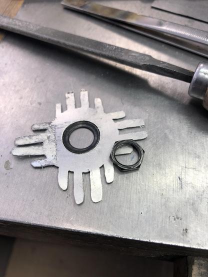 An assortment of metal tools and parts on a work surface, including a circular metal piece with grid design, a fitted black ring and a black hex nut. There are also metal files and other tools in the background.