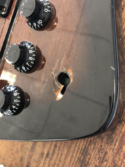 A close-up of a guitar's top, showing several black control knobs and a noticeable hole in the wood with a crack surrounding it. The surface appears glossy and polished.