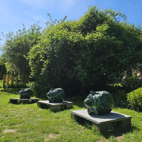 Drie glanzende, donkergroene marmeren beelden van hoofdvormen liggen op lage sokkels in een zonnige tuin met gras en omringende struiken. De hoofden kijken allemaal omhoog, en er is een helderblauwe lucht op de achtergrond.
