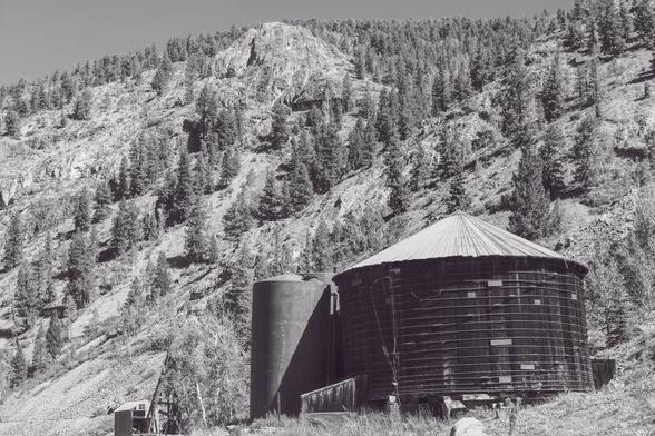 A large, weathered wooden water tank with a rusting metal roof stands near a smaller metal tank at an old mining site outside Lake City, Colorado. Towering pine-covered slopes rise steeply in the background under a clear mountain sky.