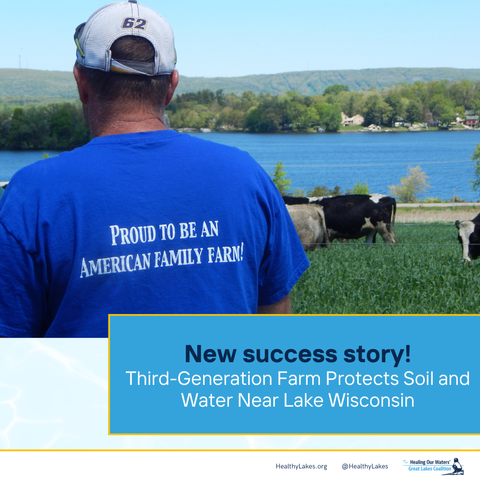 A middle-aged white man looks at a herd of dairy cows grazing on green grass next to a clear blue lake surrounded by green trees. His shirt is bright blue and reads "proud to be an American family farm!"