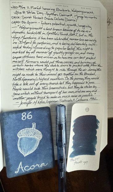 Open notebook with handwritten text from the post at the of the page with additional text: “Walpurgisnacht is best known because of its use as a dramatic backcloth in Goethe’s Faust, Part I, but in the Harz Mountains it has been celebrated, more or less seriously, on 30 April for centuries, and is being deliberately cultivated today. According to popular belief, this night is marked by all manner of ghostly goings-on, and many superstitions have arisen as to how one can best protect oneself. Farmers would put three crosses and bunches of certain herbs above the stable doors to protect their cattle from the witches which were thought to ride through the air during this night en route to their annual get-together on the Brocken, North Germany’s highest mountain. On their journey they would bite a piece out of every church bell they happened to pass. People would hide their broomsticks lest they be stolen by some witch without transport of her own, and one way and another people tried to make as much noise as possible.” — Jennifer M. Russ, German Festivals & Customs 1982. Below the text is a card with a blue on blue line drawing of an acorn. Next to the card is a clear blue plastic fountain pen and swatch of a medium blue ink with faint red sheen. 