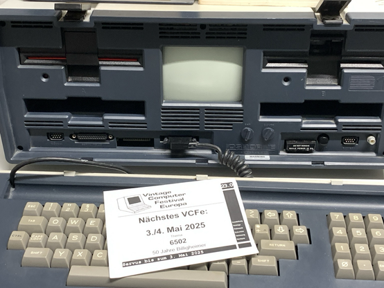 Frontalansicht eines geöffneten Osborne 1 Portable Computers mit eingebautem Bildschirm, Tastatur und Anschlüssen. Auf der Tastatur liegt ein Flyer für das Vintage Computer Festival Europa am 3./4. Mai 2025 mit dem Thema ‘50 Jahre 6502’