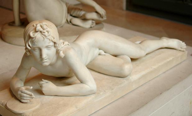 a sculpture,  very lifelike,  of a teenage boy with curly hair, he is laying on his stomach, his elbows lifting up his head and shoulders, his legs on top of each other, done in white marble 