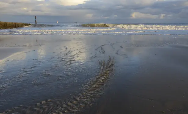 Man sieht einen braunen Strand, an dem in Rillen das Meerwasser zurück läuft. In der Ferne brechen mit weißen Schaumkronen Wellen an den Strand und am Himmel sind Wolkengebirge unterwegs.