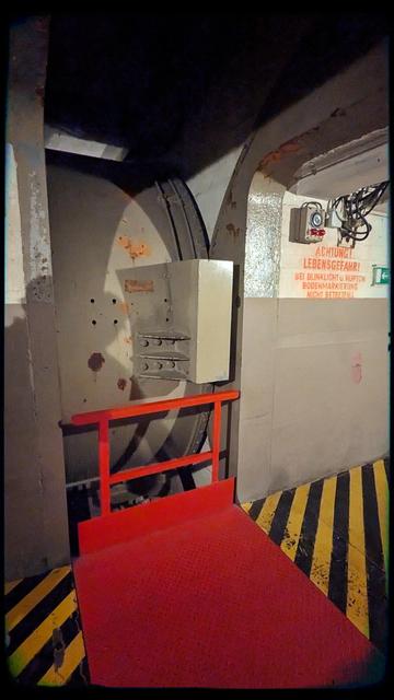 Side view of a huge, heavy vault door, currently in its opened state, embedded in the wall. There's a red footbridge part that slides out of the way when the door closes. There are black and yellow warning stripes on the floor on both sides. The walls are a very typical bunker concrete grey.