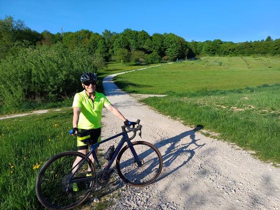 I'm standing with my Canyon Endurace along a green meadow on a gravel road near Bremthal, Hesse, Germany. It's a beautiful late spring afternoon and about 25° Celsius. I'm wearing a phosphorescent-green "kit." My bike is deep purple (it looks black here). There is a grove of low-growth trees behind me and to my right. My angular late-afternoon shadow across the path adds an interesting touch to the photo. The photo was taken by a passerby, a man hiking with his female partner.