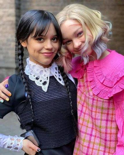 Jenny Ortega as Wednesday Adams in her black clothes and Emma Myers as Enid Sinclair in her rosa outfit. Both stand next to each other, Enid has her arm around Wednesday's shoulder and they smile into the camera.