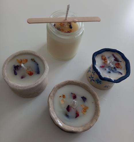 Fotografía a 4 velas. La mayor de ellas está dentro de un recipiente de cristal, con un palo con un agujero en la mecha porque se está  endurenciendo. Las tres restantes están contenidas en recipientes hechos en pasta de modelar, pintados a mano y con resina cristal. Dos de ellos tienen un granulado con café y otro está pintado con flores azules y un borde del mismo color. Todas las velas tienen en su superficie pétalos de rosas, jazmines y malvas.