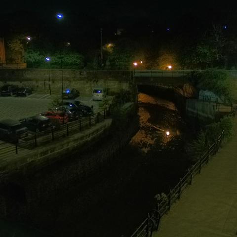 Photograph of the tidal stretch of Ouseburn River at south side of Ouseburn Bridge in Newcastle upon Tyne. To the left (south-west) of the bridge is Cut Bank, and to the right (north-east) is Byker Bank. In the foreground is the pedestrian Riverside Walk and the photo also spans some surrounding built-up areas and trees, with sky in the background. Still image captured from webcam at 03:12 hrs on Thursday 1 May 2025.