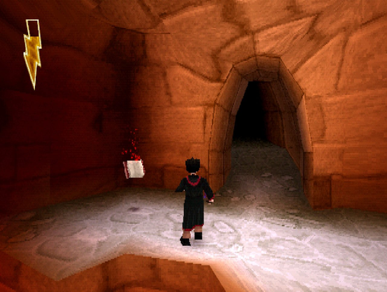 A imagem mostra uma cena de um videogame em 3D (Harry Potter and the Sorcerer's Stone de Playstation 1) com gráficos de estilo retrô. O personagem, Harry Potter, com cabelo preto e vestindo uma capa preta com detalhes vermelhos, está de costas, caminhando em direção a um túnel escuro à direita. O cenário é uma caverna com paredes e chão de pedra marrom. O chão parece coberto por uma fina camada de gelo ou neve, com tons de branco e cinza. À esquerda, há um item flutuante: um livro aberto com capa vermelhos, cercada por pequenas partículas vermelhas brilhantes. No canto superior esquerdo da tela, há um ícone de um raio amarelo dentro de um retângulo branco, indicando a energia. A iluminação é suave, com sombras escuras no túnel, criando um contraste com as áreas mais claras da caverna.