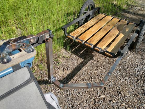 A view of a trailer hitch made out of square steel tubing welded to plate steel flames and bolted to the top of a spicy curry cargo deck in the lower left foreground, with the hitched up trailer in the upper right of the frame. The towing arm of the trailer is also bare steel, roughly welded out of several sections of square tube, while the trailer frame is black painted tubing with a deck of 5 pallet wood planks, sanded, linseed oiled, and lashed to the frame with strips of old inner tube.