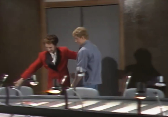 A imagem mostra Odete Roitman, interpretada pela atriz Beatriz Segall na novela Vale Tudo (1988), vestindo um blazer vermelho e um top branco, com um ar de intriga ou surpresa em seu rosto. Ela está inclinada sobre uma mesa com vários microfones, aparentemente em um estúdio de televisão ou rádio. Ao seu lado, um homem de camisa azul claro observa-a. O cenário é discreto, com tons neutros e uma atmosfera que sugere um ambiente profissional e talvez tenso. A imagem parece ser um still de uma cena da novela, capturando um momento de interação entre os personagens. A qualidade da imagem é um pouco baixa, mas os detalhes principais são visíveis.