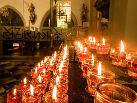 Das Bild zeigt eine lange Reihe von brennenden Kerzen in einem Innenraum, in der Pfarrkirche in Leibnitz. Die Kerzen stehen dicht nebeneinander auf dunklen, metallenen Ablageflächen, in mehreren parallelen Reihen.

Jede Kerze befindet sich in einem kleinen, durchsichtigen Glasbehälter. Die Flammen der Kerzen sind hell und flackern leicht. Sie werfen ein warmes, gelbliches Licht auf ihre Umgebung.

Im Hintergrund ist ein Teil der Kirche mit Statuen und Bögen zu erkennen.

Der Boden im Vordergrund ist gefliest. Die Perspektive des Bildes ist leicht erhöht, sodass man gut über die Reihen der Kerzen blicken kann. Die Atmosphäre wirkt ruhig und andächtig, geprägt vom sanften Schein der vielen kleinen Flammen.