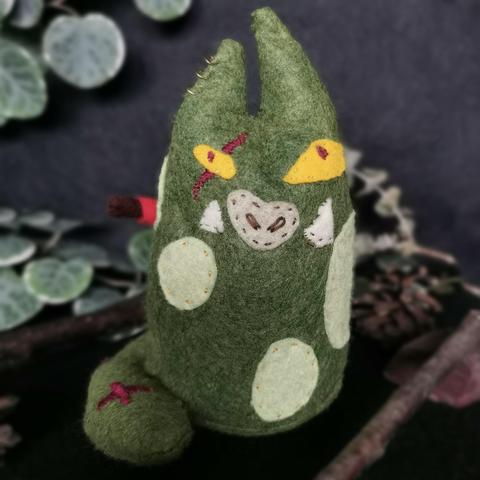 A plushie sitting on a dark backdrop with decorative plants. The plushie is of a car-like orc with mossy green skin and pale green spots. It has uneven yellow eyes with red pupils and a red scar embroidered over it’s right eye and the tip of it’s tail. 
It’s nose is snout_like and it has two tusks on each side of it. 
Three small ring piercings decorate it’s right ear. 