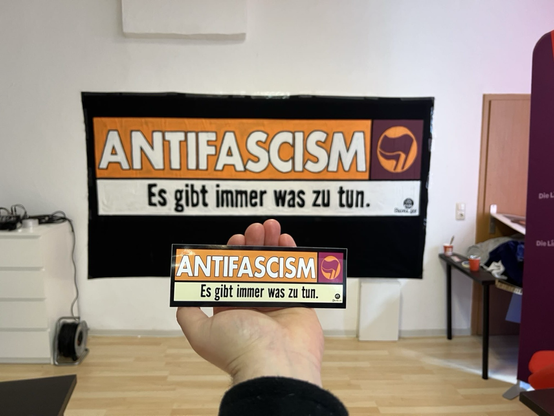 Eine Hand hält einen Aufkleber „Antifascism. Es gibt immer was zu tun“ dahinter hängt ein Transpi im exakten Stil des Aufklebers an der wand. Beides ist nach einer Baumarkt-Werbung gestaltet.