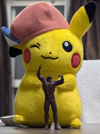 Closeup of a small, all brown figurine of a muscular male body with the head of a mole poses in front of a standing up pikachu pokemon plush that's about double the height. It's wearing a pink baseball hat sideways, which hides the right ear and only shows the pointing up left ear. This was taken in a hotel room in Osaka, Japan. Unfortunately they had to separate at the end of the trip, so Pikachu moved to germany living a peaceful life, while the Mole Man now resides in the US, pursuing his bodybuilding career.