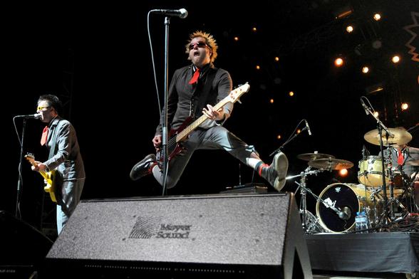 Contre-plongée. On est a un concert de Toy Dolls. Les jambes écartées, le bassiste saute au-dessus d’un retour. À gauche, on voit le chanteur avec sa guitare, près du micro, et à droite, la batterie.