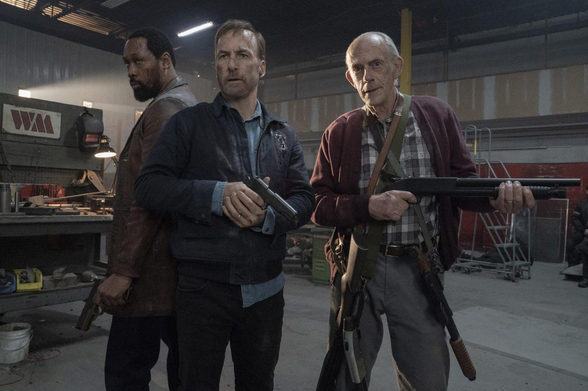 A imagem mostra três homens em uma oficina ou armazém industrial, armados e em uma postura tensa. No centro está Hutch Mansell (Bob Odenkirk), com uma jaqueta jeans escura e uma pistola na mão. À sua esquerda está Harry Mansell (RZA), também armado com uma pistola. À direita de Hutch está David Mansell (Christopher Lloyd), um homem mais velho com um casaco xadrez e uma espingarda de cano curto, carregando outras armas em uma bandoleira. O fundo é escuro e industrial, com máquinas e equipamentos visíveis, criando uma atmosfera sombria e de confronto. A iluminação destaca os três homens contra o ambiente escuro. A cena, do filme Nobody, sugere uma situação de confronto ou preparação para uma ação violenta.