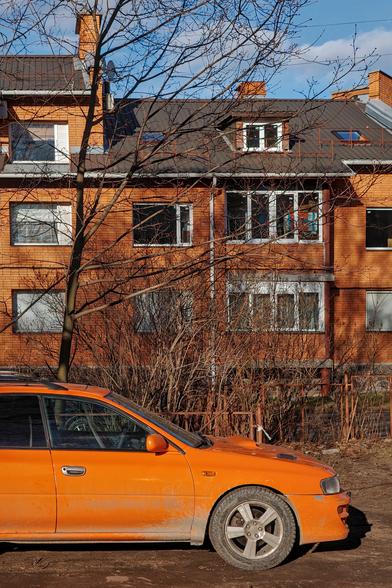 An orange car stands in the foreground. In the far background is a red-orange house. Above the house is a blue sky.
