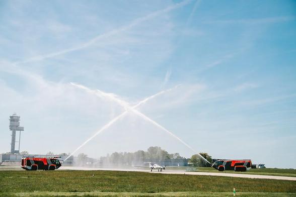 Sprühbogen der Flughafenfeuerwehr zur Begrüßung der SEABIRD 3: 
2 Feuerwehrfahrzeuge links und rechts einer Rollbahn rahmen das Flugzeug in ihrer Mitte mit zwei sich kreuzenden Sprühstrahlen ein. Im Hintergrund ein Flughafentower.