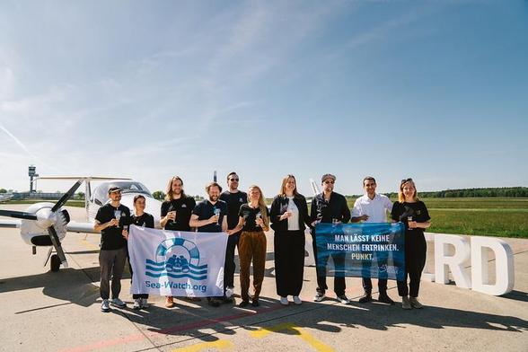 Menschen mit zwei Bannern "Sea-Watch.org" & "Man lässt keine Menschen ertrinken. Punkt. United4Rescue" vor der SEABIRD 3 auf dem Rollfeld. Sie halten Sektgläser in der Hand. Auf ihren Shirts Logos von Sea Eatch, HPI Suisse & United4Rescue.