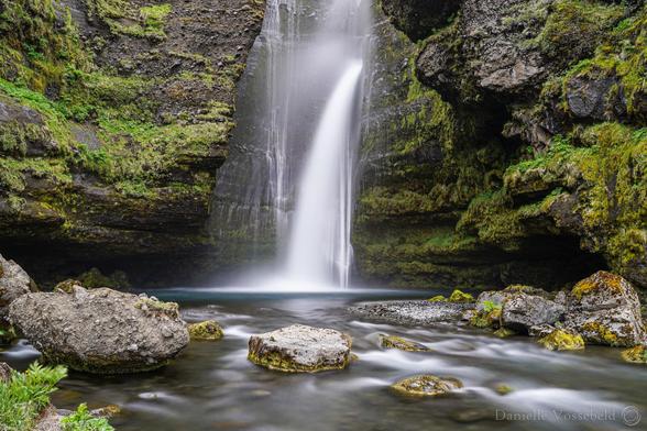 In an inlet in the rocks, a bright white stream of water flows down. The water has a little turquoise pool, before it flows towards the photographer (yep, me). There is moss on the walls of the rocks, there are boulders in the stream. 