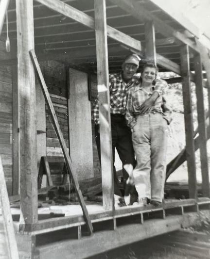 A man with his arm around a woman’s shoulder. A woman in jeans and a plaid shirt. They are standing on an unfinished porch. 