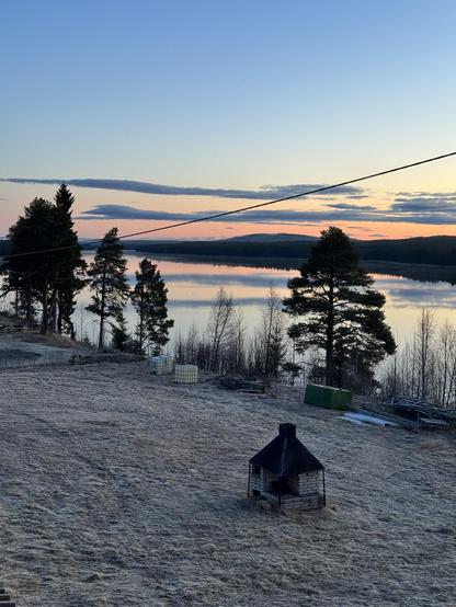 Frost-covered yard by the Vindelälven river in Swedish Lapland at 4 am sunrise, with a brick smoker in the foreground and still water reflecting the glowing sky and pine trees.