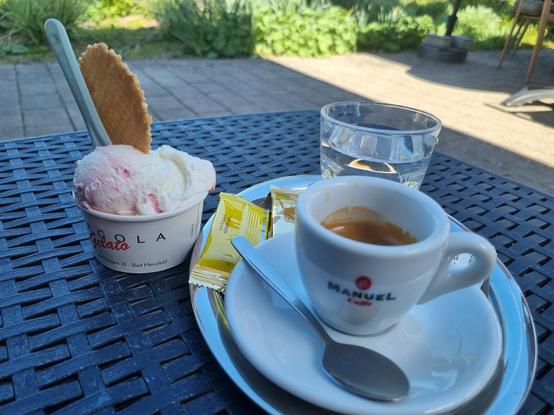Eine Kugel Amarena-Eis im Pappbecher und ein Tablett mit Espresso, Wasserglas und Keksen auf einem Hocker aus Kunststoffgeflecht. im Hintergrund sind gepflegte Rabatten eines Kurparks zu erahnen.