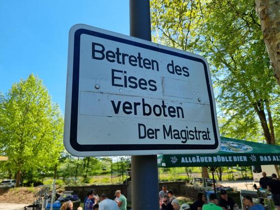 Ein Schild mit der Aufschrift "Betreten des Eises verboten -- Der Magistrat". Dahinter sitzen Leute in einer Art Biergarten und löschen ihren Durst.