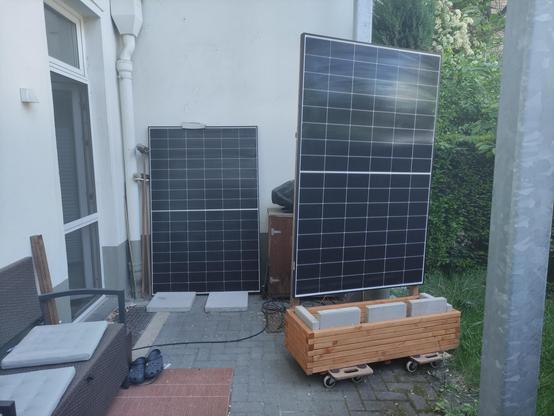 Zwei senkrecht stehenden Solarpanels auf einer Terrasse: Eines lehnt an der Wand, das andere ist auf einen rollenden Blumenkübel montiert.
