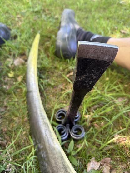 The image shows a close-up of a curved scythe blade and a black metal peening anvil with decorative spirals, resting on grass. A foot wearing a sneaker is partially visible in the background.