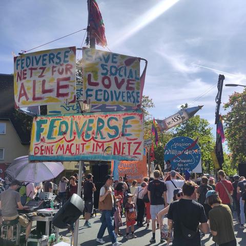 Auf einer 1. Mai demo im Grunewald steht ein Schild mit der Aufschrift: " Fediverse, Netz für ALLE, save social love, Fediverse, deine Macht im Netz" Im Hintergrund eine Bühne.