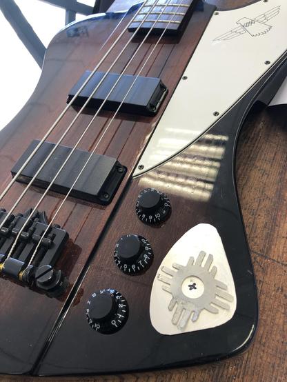 Close-up of a bass guitar featuring a wood body with four strings and black pickups. A triangular silver cover with a circular grid design is visible on the lower part of the guitar, along with a white pickguard design. The background is blurred, focusing on the guitar's features.