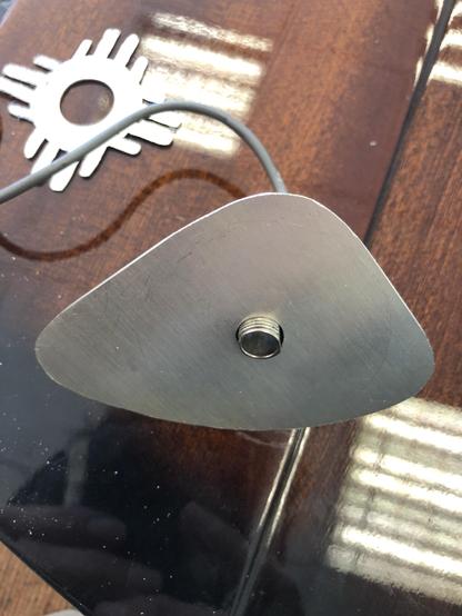 A close-up of a metallic, triangular object with a smooth surface and a threaded hole at its center, resting on a wooden table. A gray cable is visible attached to the object. The background features reflections and a circular design.