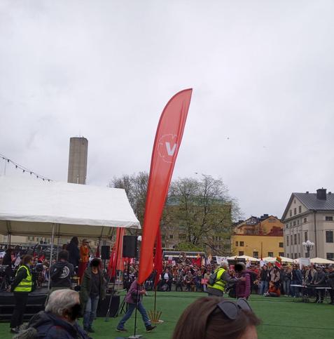 A square with a temporary scene with people on and around it. Houses in the background. In the middle a red banner with the symbol of Vänsterpartiet which is the Swedish left wing party. 