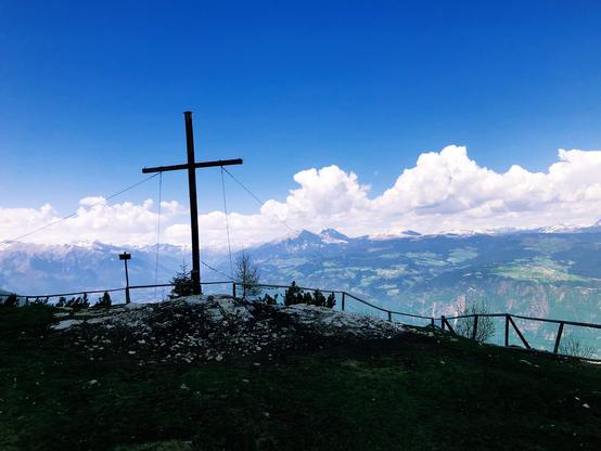 A cross at the summit of the mountain with a fence a mountain peaks in the distance