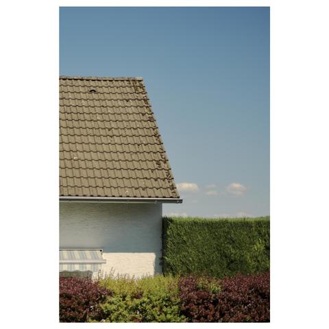 Hinter einer Hecke blickt man auf ein weißes Satteldach Haus mit halb ausgefahrener Markise.  Neben dem Haus erhebt sich eine weitere eckig geschnittene Hecke vor blauem Himmel. 