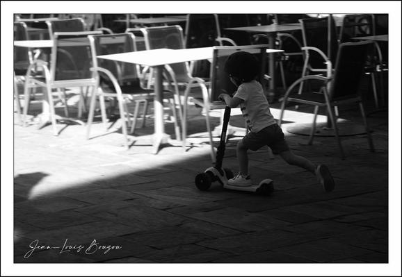 The image captures a young child energetically riding a scooter, creating a sense of movement and joy. The child is slightly blurred, emphasizing speed and playfulness, while the surrounding area is filled with empty tables and chairs, suggesting a quiet moment in an outdoor cafe.
The contrast between the light and shadow adds depth, highlighting the child's figure against the darker background. The use of black and white intensifies the visual drama, focusing the viewer's attention on the child and their joyous activity. This choice might evoke a sense of nostalgia or timelessness, representing carefree childhood moments.
Overall, the scene conveys a vibrant, playful energy set against a serene, almost still backdrop, emphasizing the contrast between youthful exuberance and the calm of a cafe setting.
