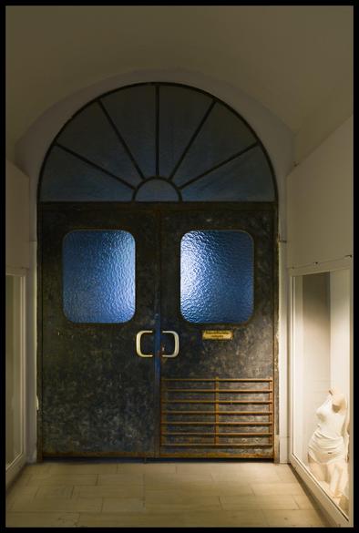 A dimly lit hallway featuring imposing, dark metal double doors set within an arched doorway. The doors have square frosted glass windows glowing with cool blue light from behind, and an arched fanlight window above. To the right, a partial mannequin torso stands in a recessed display area next to the doorway. The floor is tiled, and the overall atmosphere is mysterious and slightly industrial.