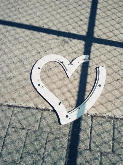 A broken white heart shaped picture frame lies on a sidewalk. The shadows from a nearby chain link fence can also be seen.