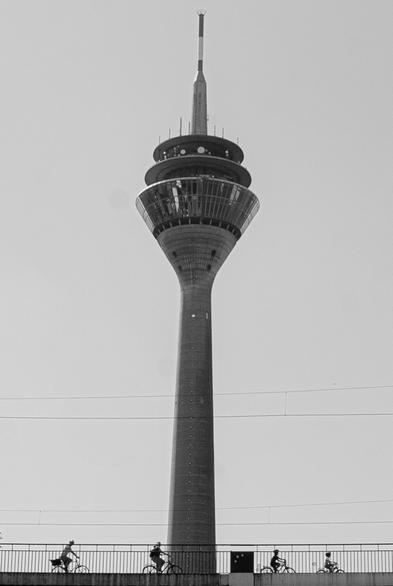 Ein hoher Turm ist prominent gegen einen klaren Himmel zu sehen, während mehrere Radfahrer auf einem Weg im Vordergrund fahren. Das Bild ist in Schwarzweiß und hebt die Struktur des Turms und die Silhouetten der Radfahrer hervor.

A tall tower is prominently featured against a clear sky, while several cyclists ride along a path in the foreground. The image is in black and white, highlighting the tower's structure and the cyclists' silhouettes.