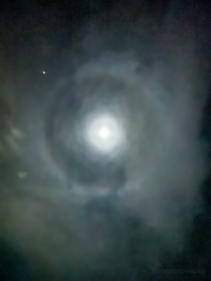 A bright full moon surrounded by a large, faint circular halo in a cloudy night sky. A bright point of light, likely a planet, appears to the upper left of the moon. The halo is caused by ice crystals in the upper atmosphere refracting the moonlight.