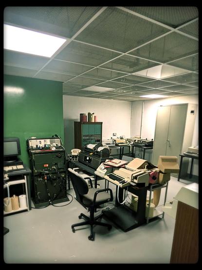 An office space in a nuclear bunker. Equipped with desks and desk chairs, a decoding machine, an old tv, rotary phone and several typing machines and (I'm assuming) super secret communication devices. Filing cabinets are spaced along the green and grey walls.