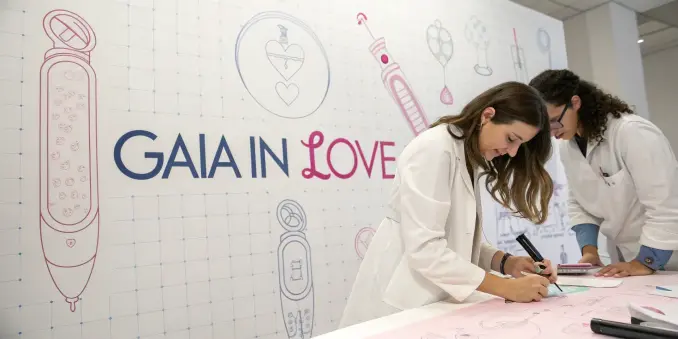 Two women in white lab coats are sketching product designs on a large desk in front of a Gaia in Love logo. The background wall is filled with blueprint-style drawings of sex tech devices, symbolizing female-led innovation in intimate wellness.